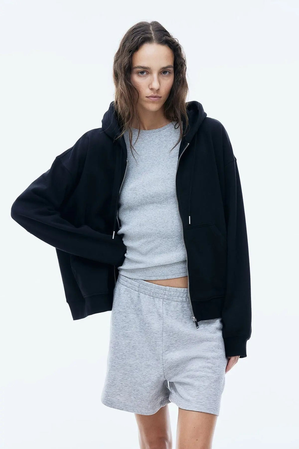 Woman wearing black zip-up hoodie over grey crop top and grey shorts, standing against white background
