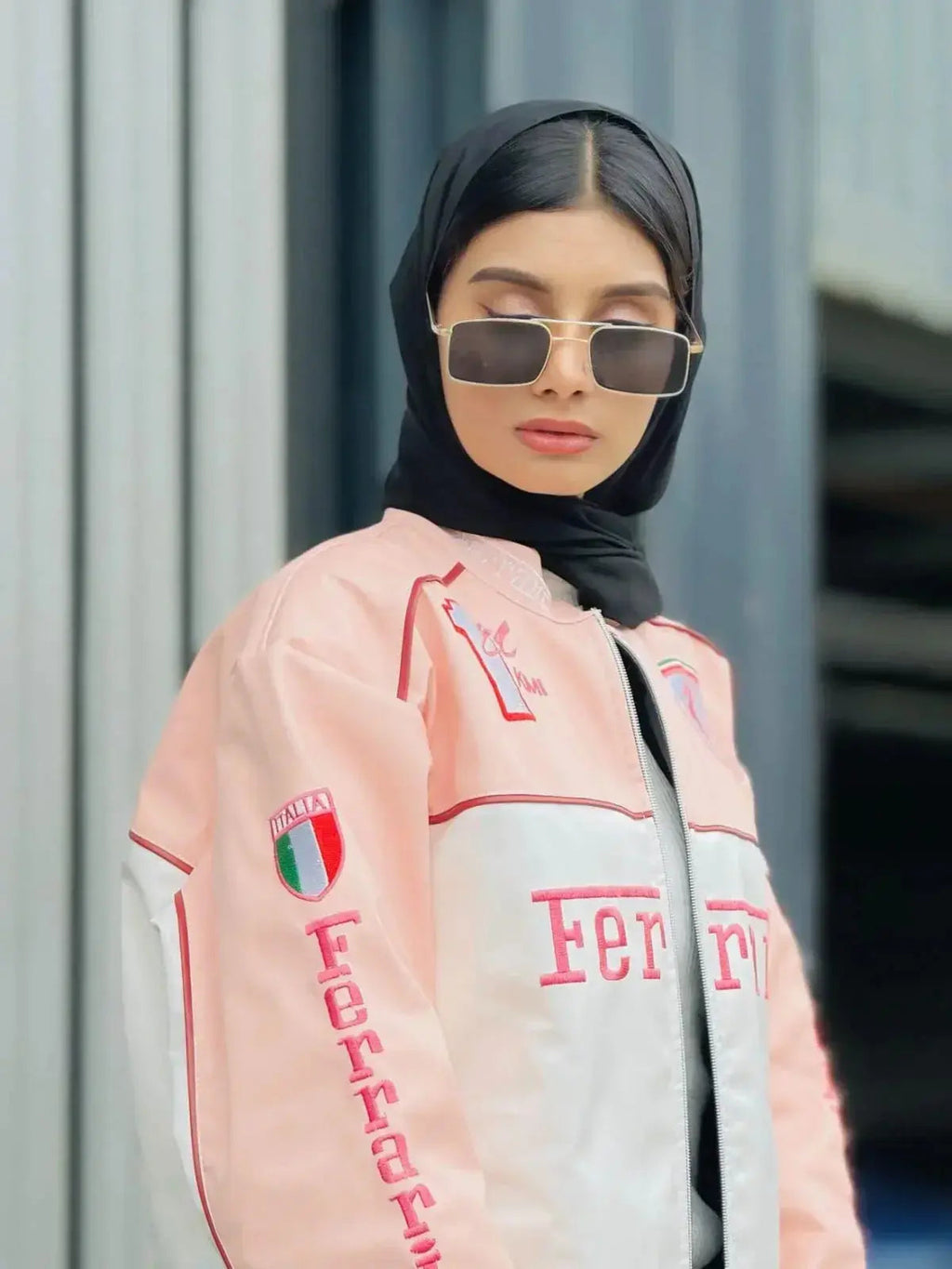 Woman wearing a pink Ferrari racing jacket, sunglasses, and a black hijab in urban setting